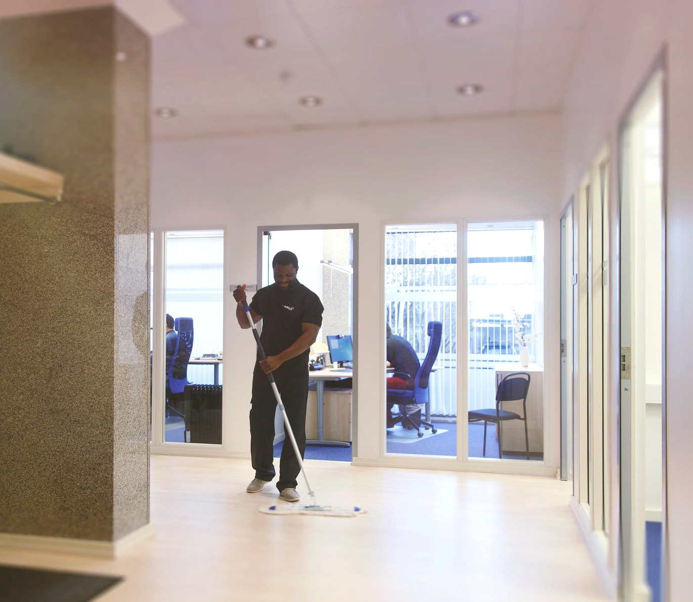 Man mopping an office floor. White walls, large windows, desks in background.