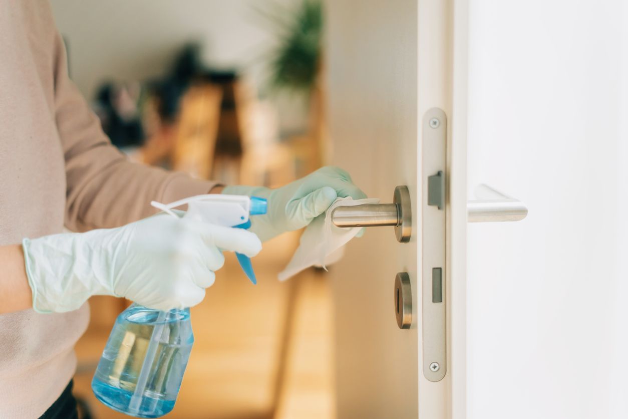 Person med hansker sprayer og tørker av et dørhåndtak med desinfeksjonsmiddel.