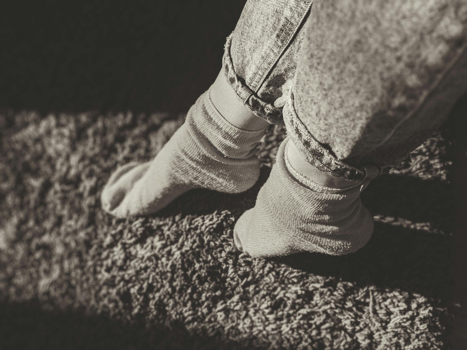 Føtter i teksturerte sokker og jeans hviler på et rufsete, mørkt teppe.