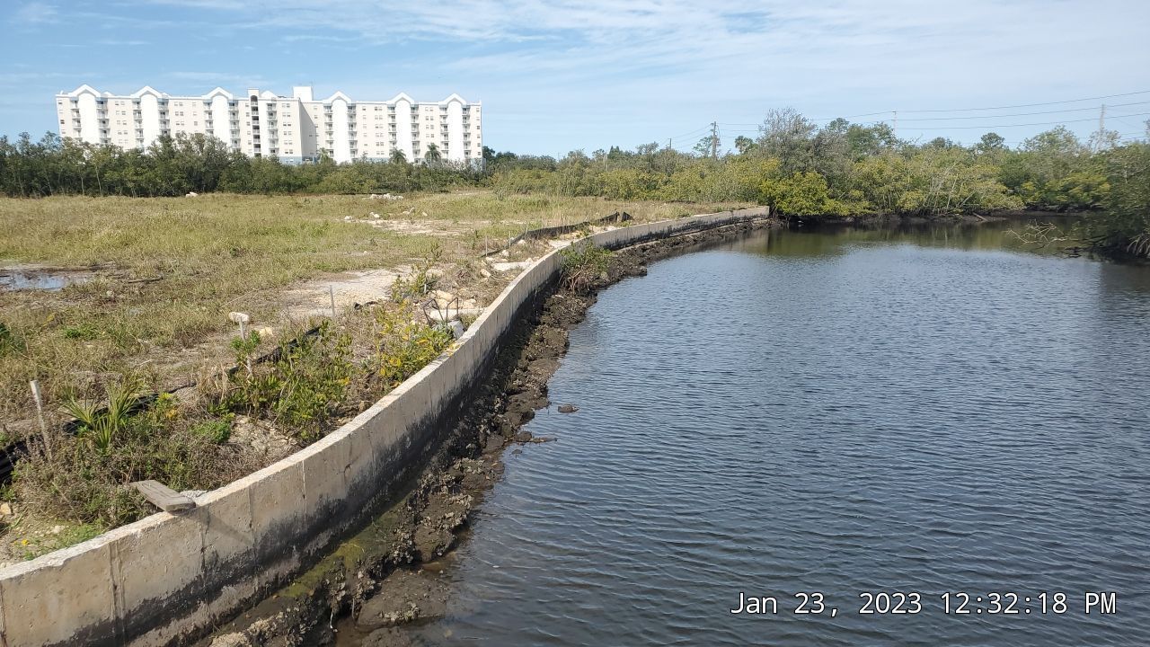 Curved concrete embankment beside a calm waterbody, with apartment buildings in the distance under a blue sky.