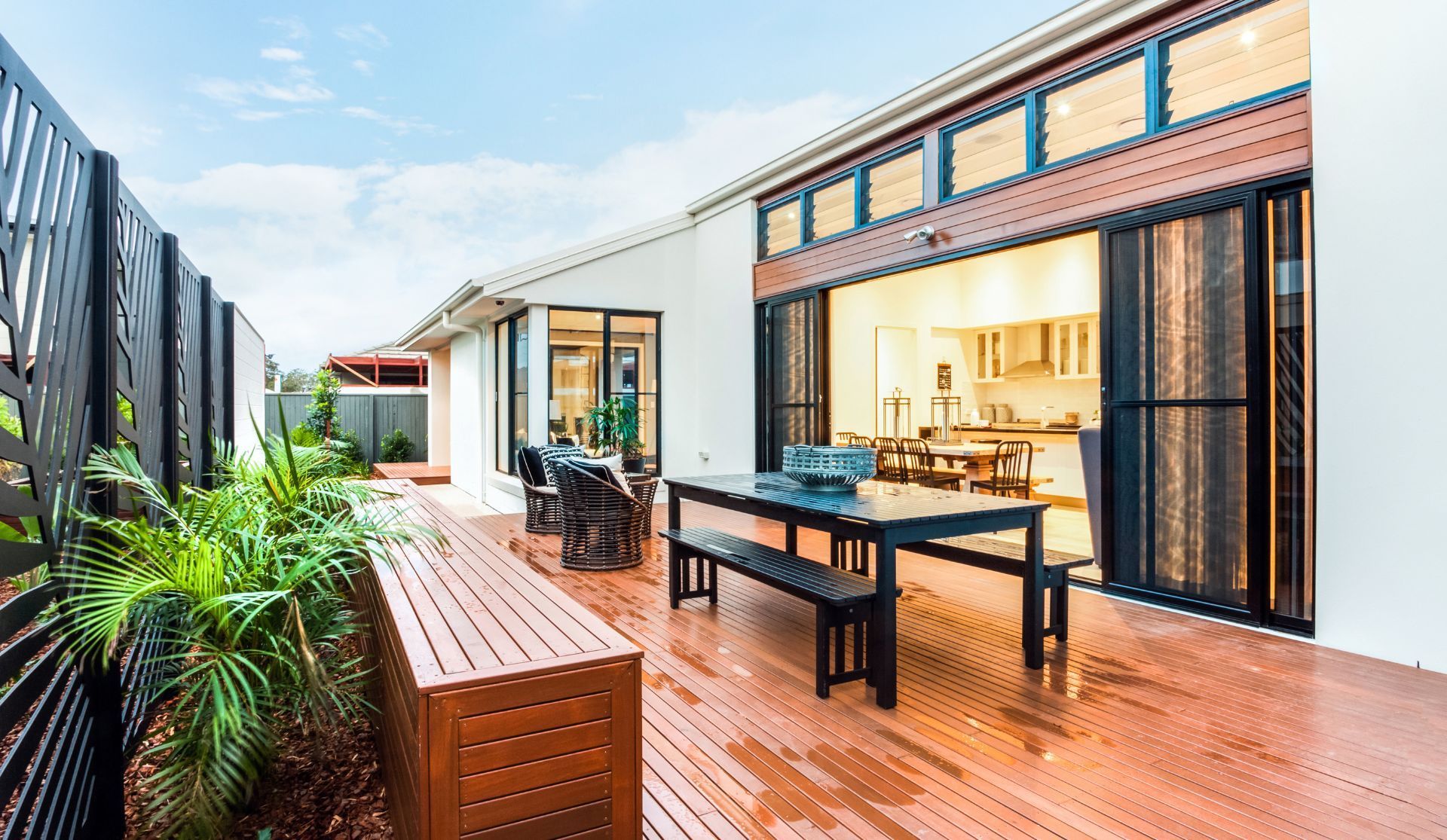 Modern patio with wooden deck, black outdoor furniture, and glass doors beside lush greenery