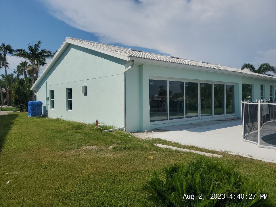 Mint-green house with large sliding glass doors, grassy yard, and palm trees under a partly cloudy sky