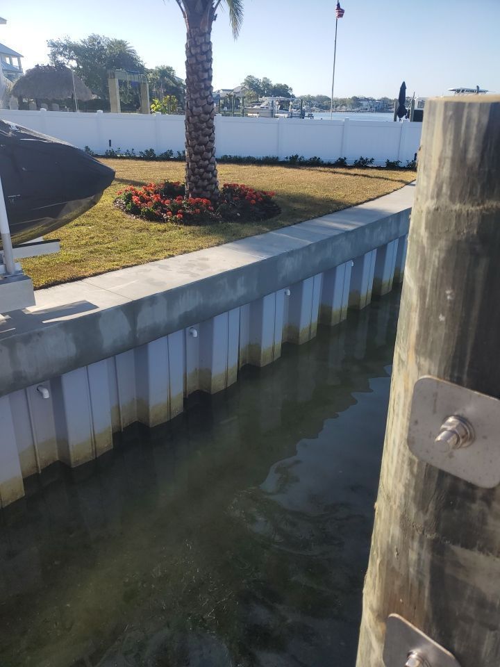 Canal beside a white waterfront wall, with palm trees, flowers, and a boat dock post in the foreground