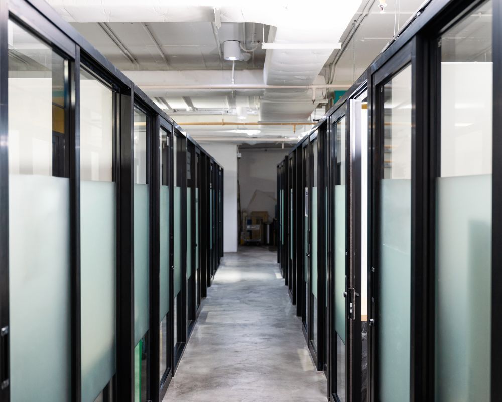 Modern office hallway lined with glass-walled rooms and black-framed doors on both sides