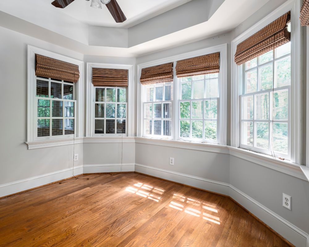 Bright bay-windowed room with hardwood floors, white walls, and woven blinds letting in sunlight