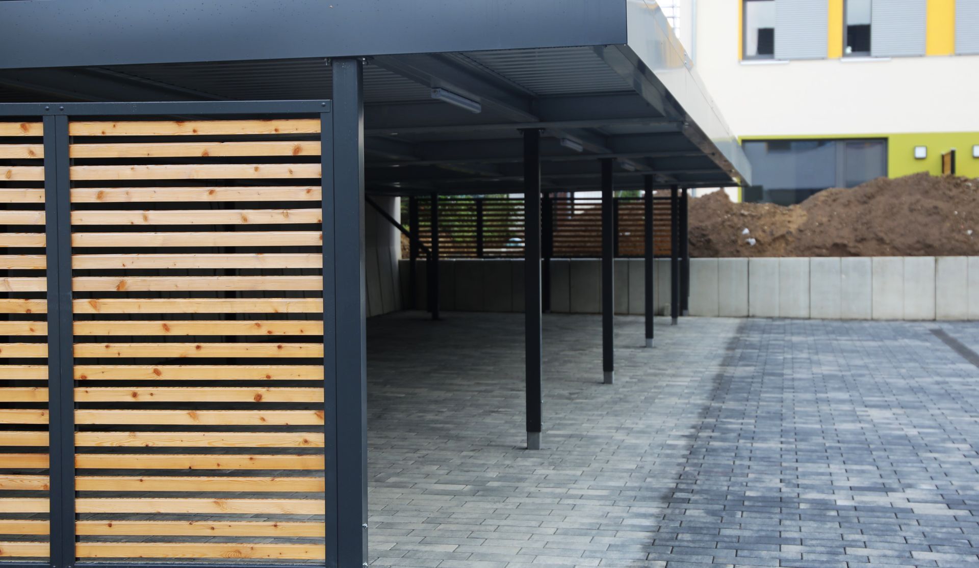 Covered parking area with black metal posts, wood slat wall, and paved ground beside a building.