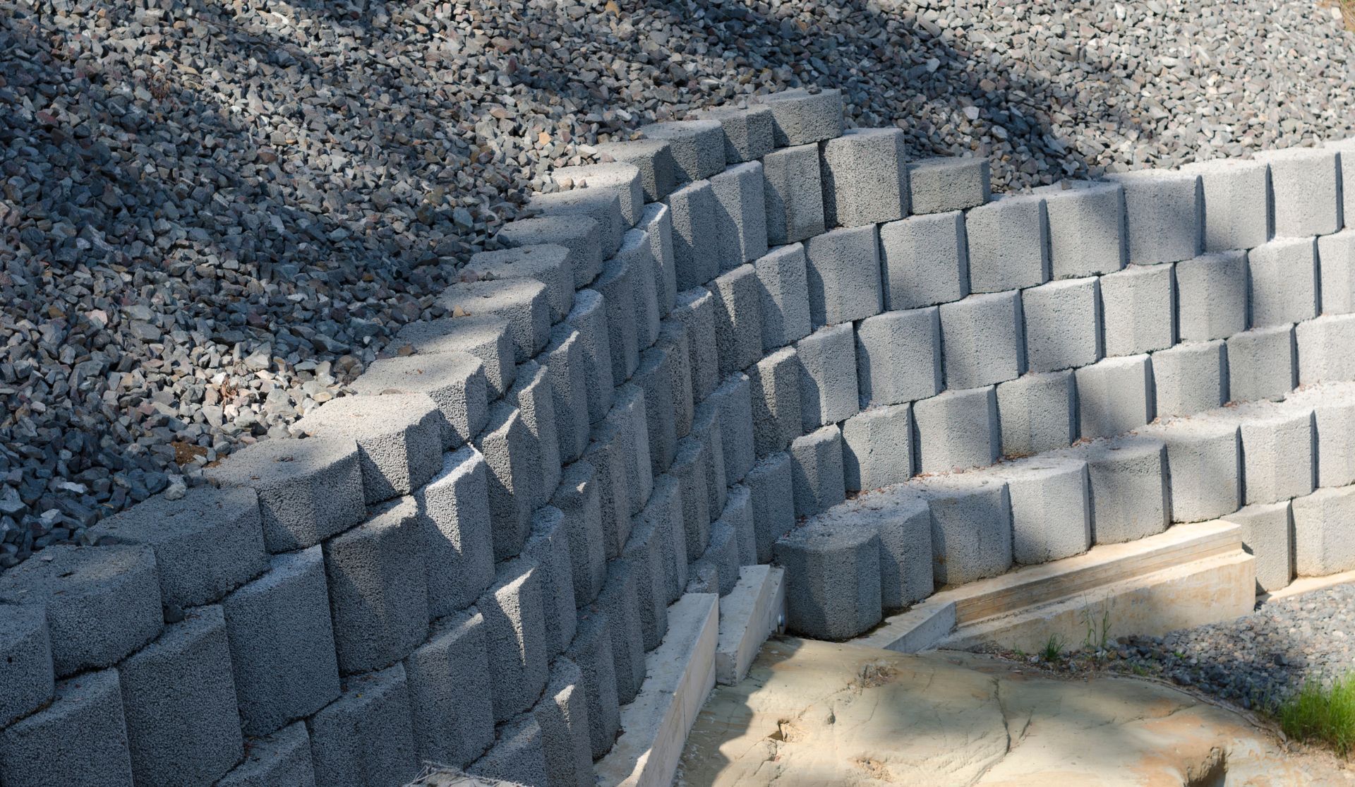 Curved gray block retaining wall built into a sloped, gravelly landscape.