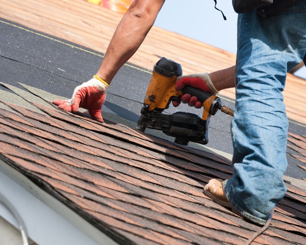 Workers installing brown shingles on a roof with a nail gun