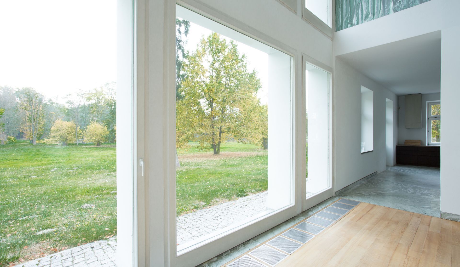 Bright modern room with floor-to-ceiling windows overlooking a green lawn and trees, wood floors inside