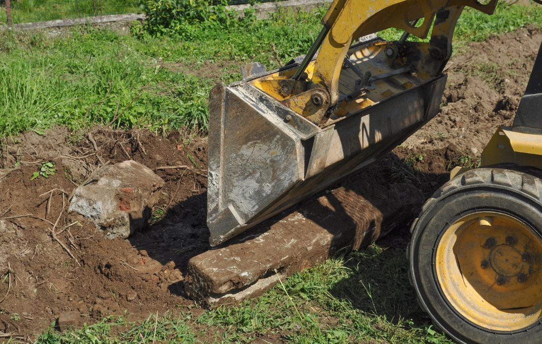 Front loader bucket digging dirt beside a buried log in a grassy yard