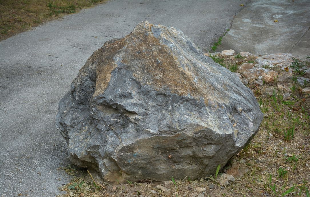 Large gray boulder on a paved path beside grass and dirt