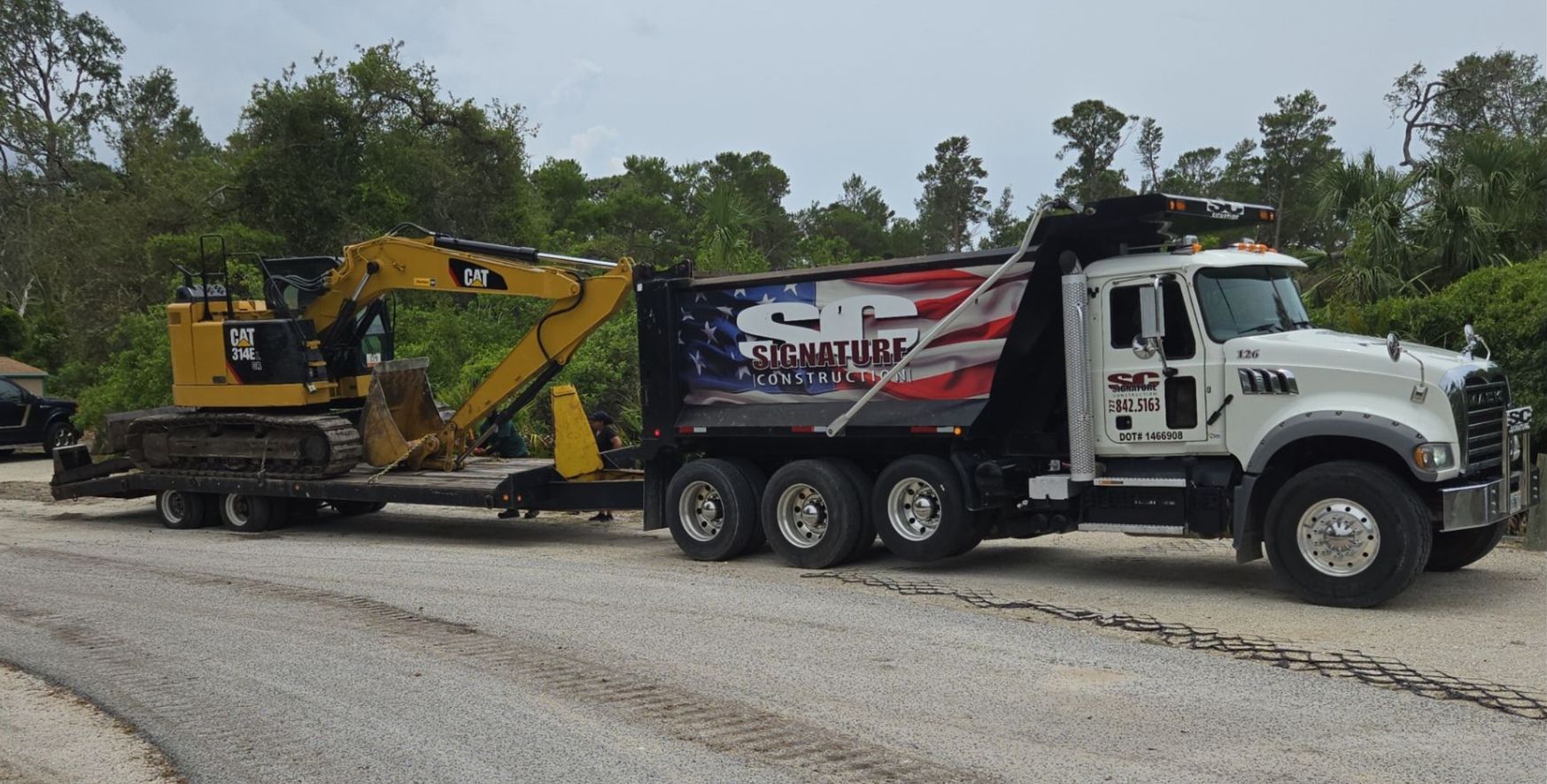White dump truck hauling a yellow excavator on a trailer along a gravel road, with trees in the background