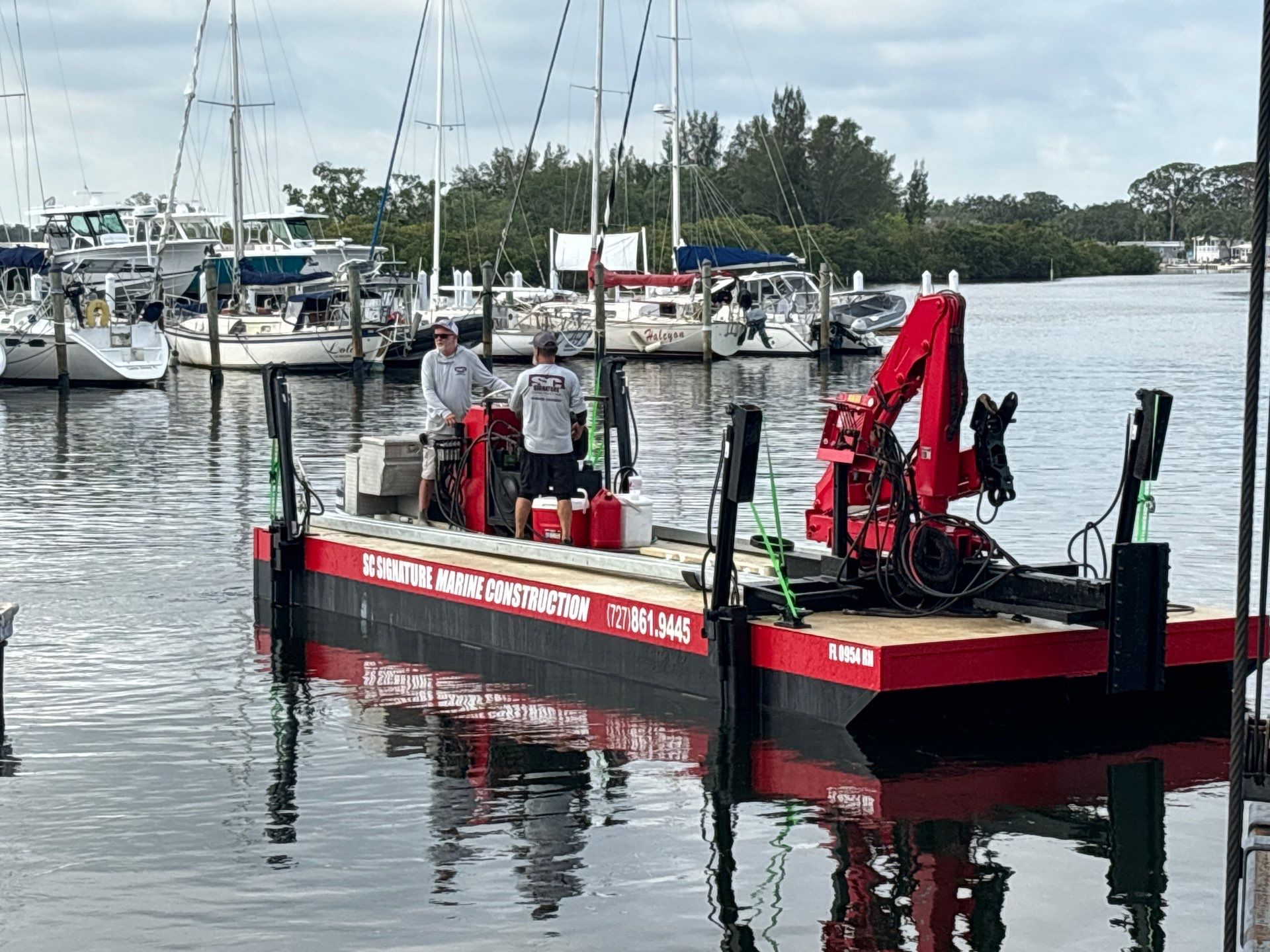 Red work barge with crane and crew docked in a marina with sailboats and calm water