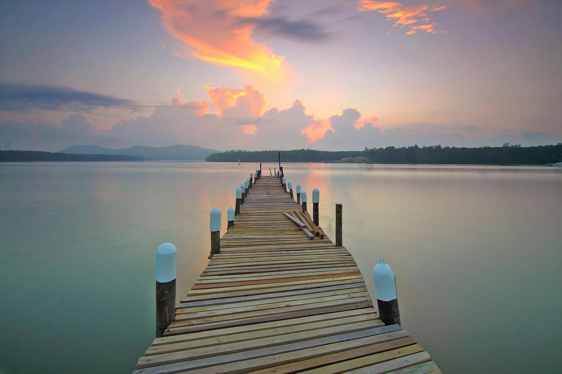 Wooden pier stretching into calm water at sunset, with pink clouds and distant mountains.