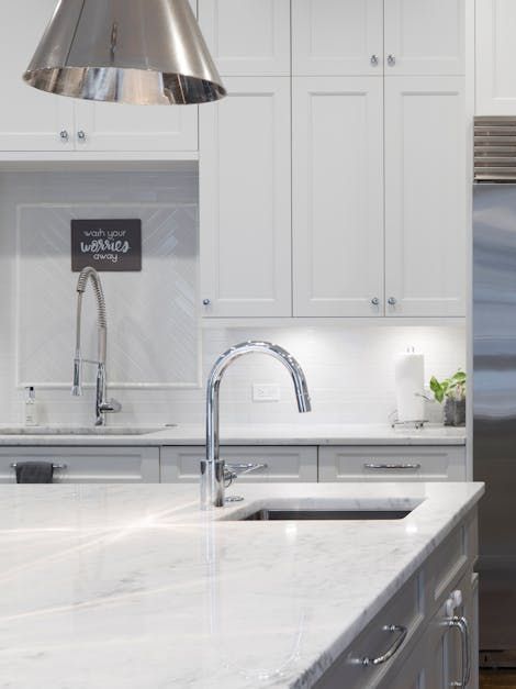 Bright white kitchen with marble island, stainless sink, and chrome faucet under pendant light.