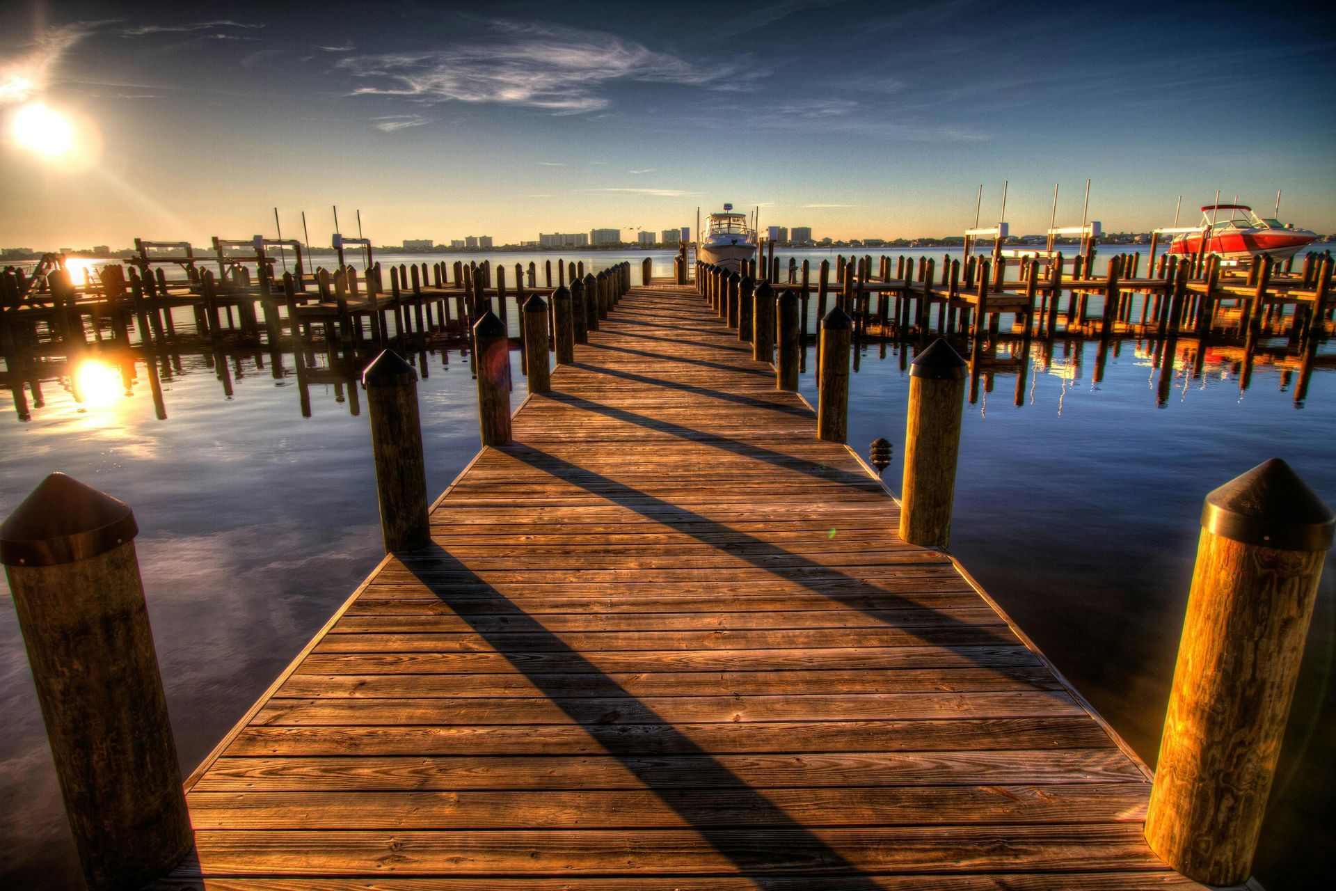 Wooden dock at sunset with long shadows stretching toward calm water and boats in the distance