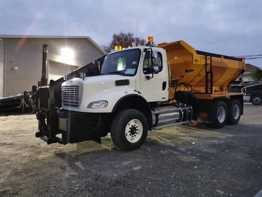 Un camion-benne blanc équipé d'un épandeur de sel orange est stationné sur un terrain gravillonné au crépuscule.