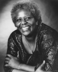Smiling woman in a shimmering top, leaning forward against a soft studio backdrop.
