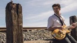 Man holding a yellow guitar beside weathered wooden posts in a rocky outdoor landscape