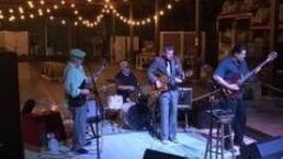 Live band performing on a small indoor stage with guitars, drums, and stools under warm lighting