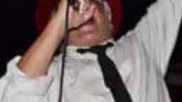 Person singing into a microphone with one arm raised, wearing a white shirt and black tie against a dark background