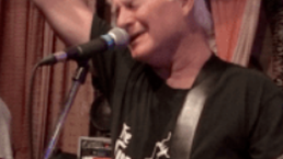 Singer at a microphone, smiling onstage with a guitar strap over a black shirt.