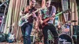 Two musicians playing electric guitars on stage in front of drums and tall wooden backdrop