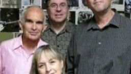 Four people pose closely together indoors, with framed photos visible in the background.