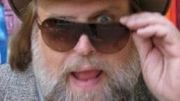 Close-up of a bearded person adjusting dark sunglasses, tongue out, wearing a hat