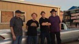Four people standing beside a silver car in front of a wooden building at dusk.