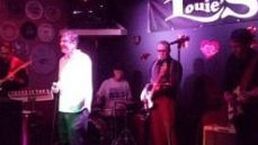 Small live band performing on a dim purple-lit stage with microphones, guitar, and drums.