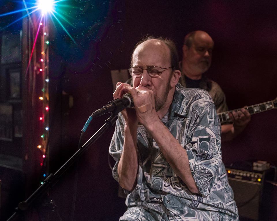 Singer playing harmonica into a microphone on a dim stage with a guitarist in the background
