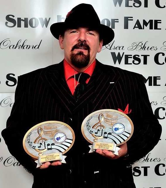 Man in black suit and fedora holds two award plaques at a Western Hall of Fame backdrop