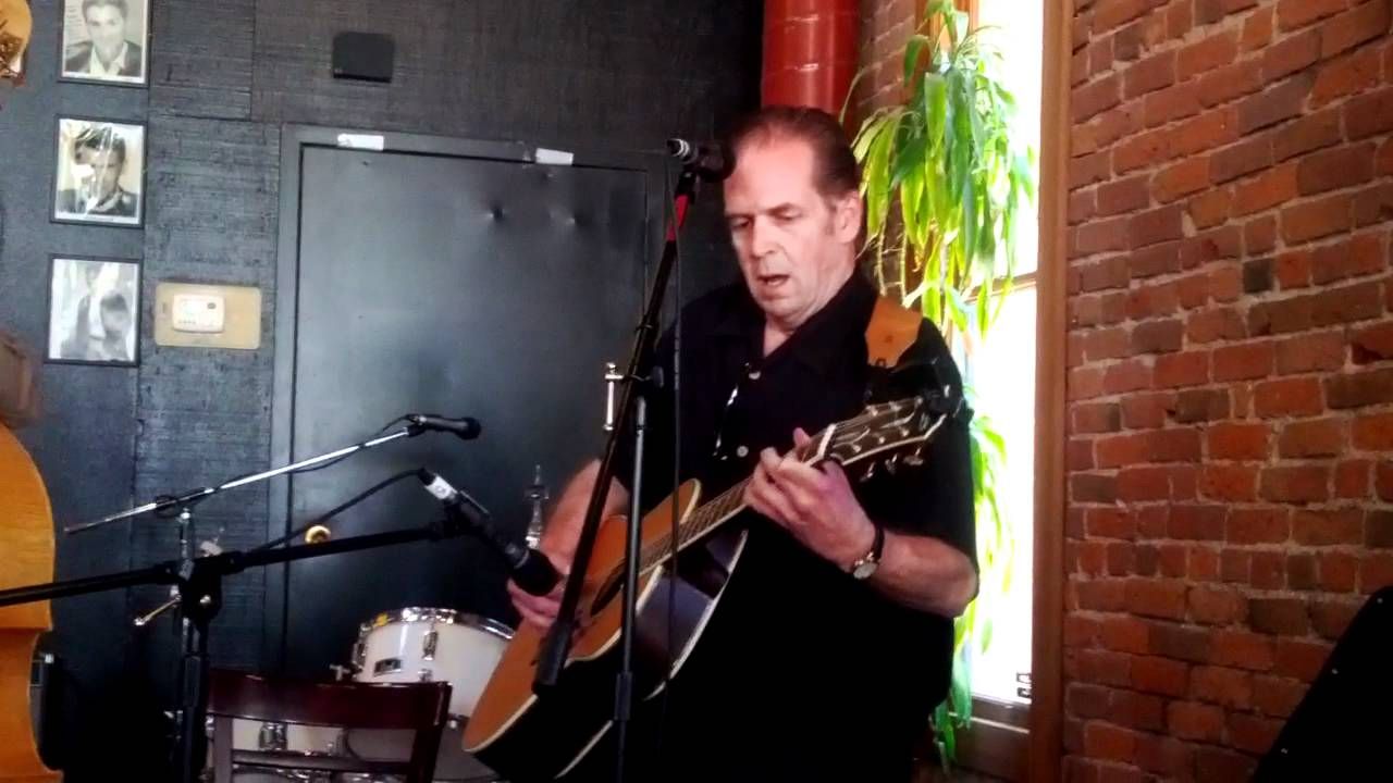 Musician singing and playing acoustic guitar on a small stage with brick wall backdrop