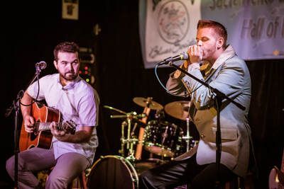 Two musicians perform onstage, one singing into a microphone and one playing acoustic guitar, with drums behind them