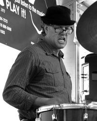 Man wearing a black hat plays drums, mouth open, in a black-and-white performance scene