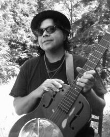 Person in sunglasses and hat holding a guitar outdoors, with trees in the background