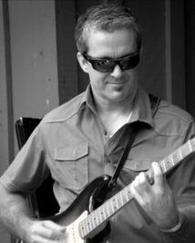 Man wearing sunglasses plays an electric guitar outdoors in a black-and-white photo