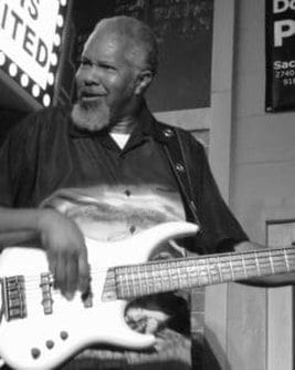 Smiling man playing a white bass guitar onstage in black and white.