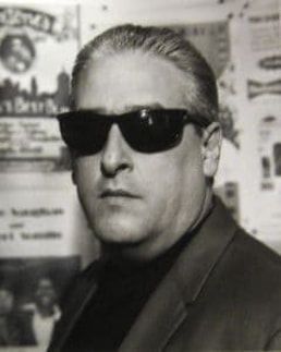 Man in dark sunglasses and suit jacket posing against a poster-covered wall