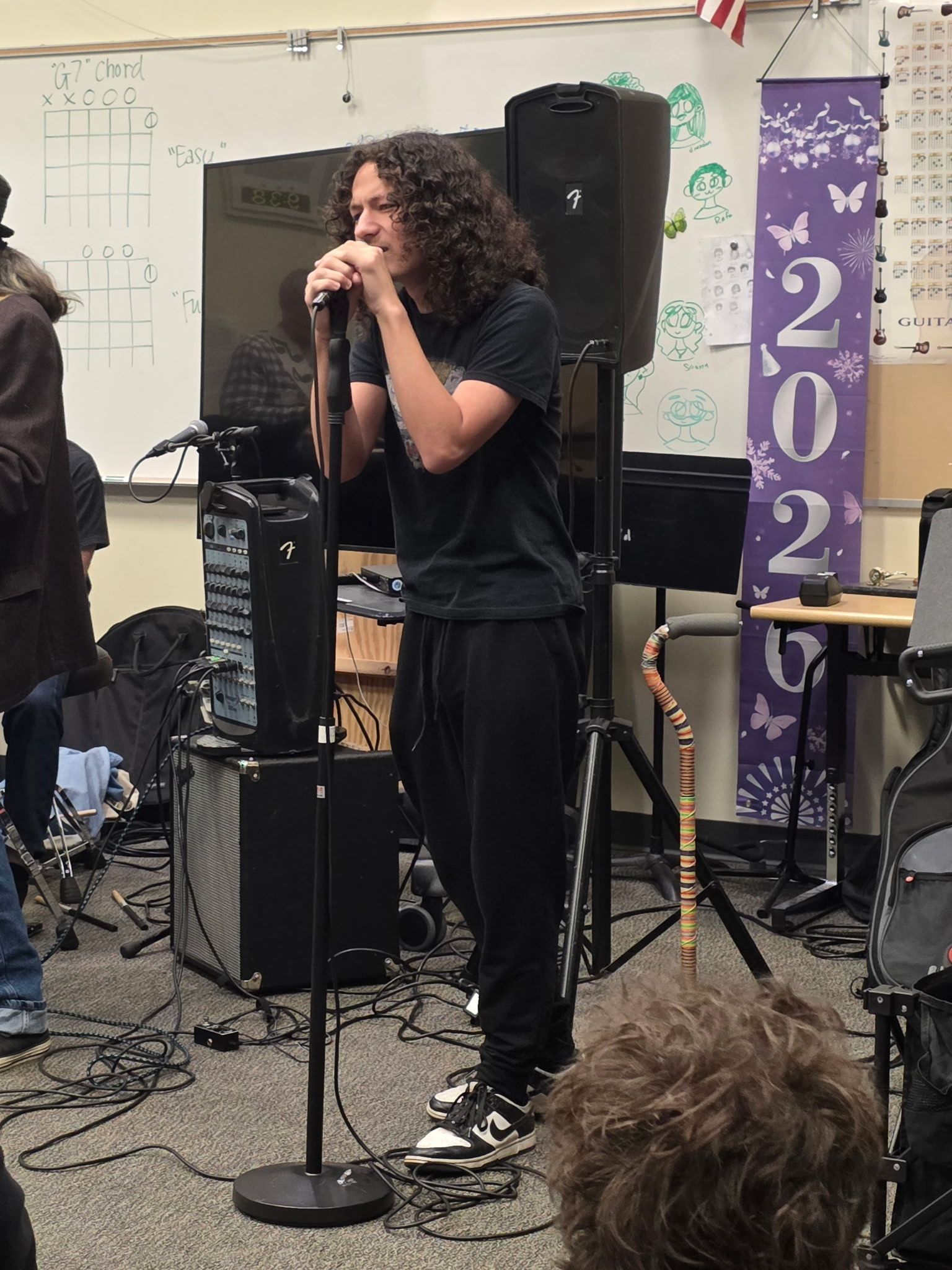 Person singing into a microphone on a small stage with speakers and a purple banner behind them.