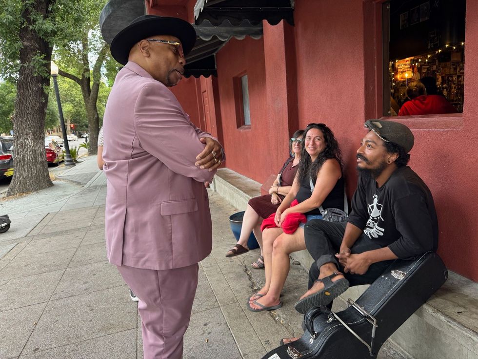 Man in pink suit and cowboy hat talks to two seated people outside a red building.