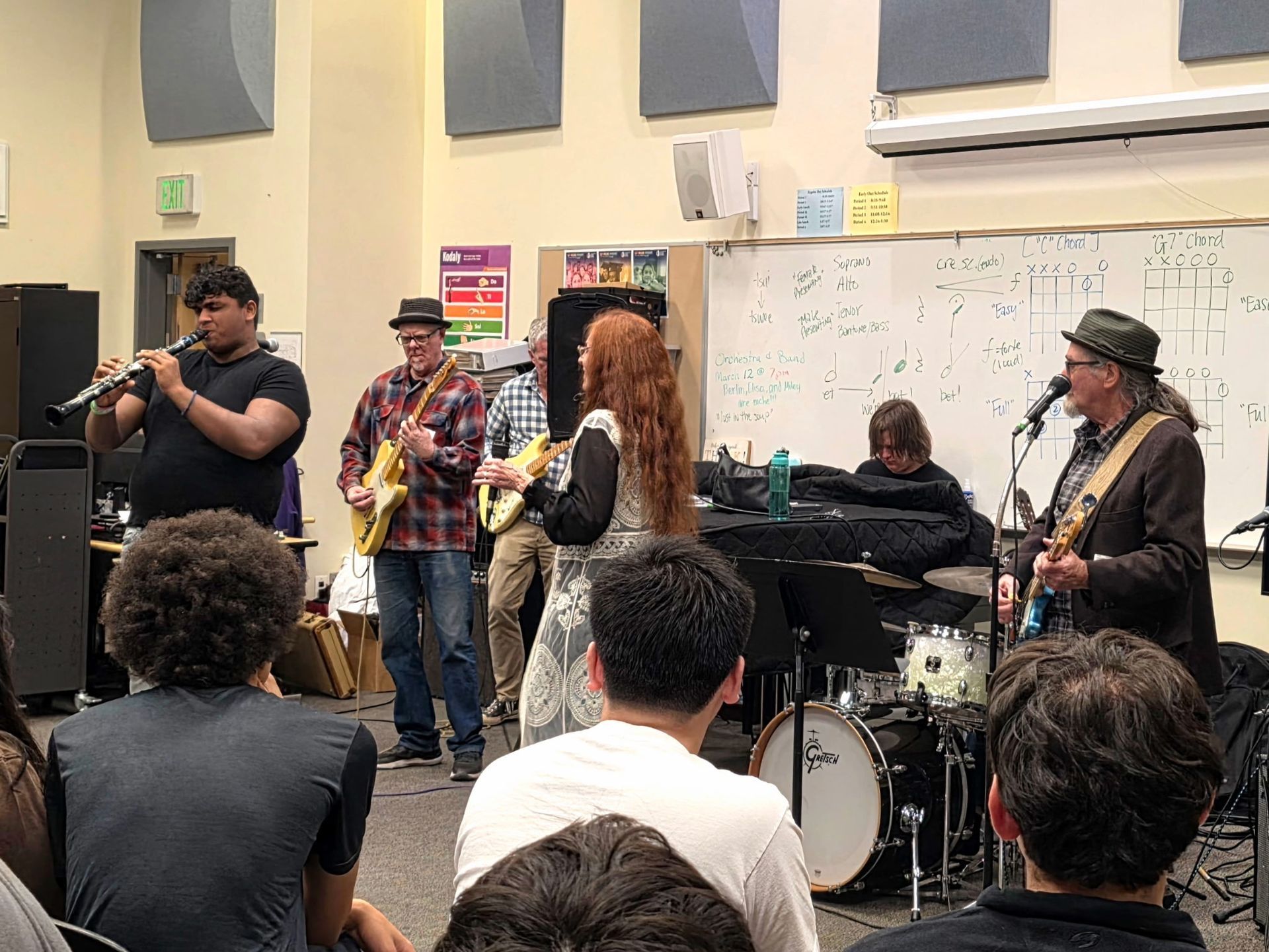 Live band performing in a rehearsal room, with audience seated in the foreground.