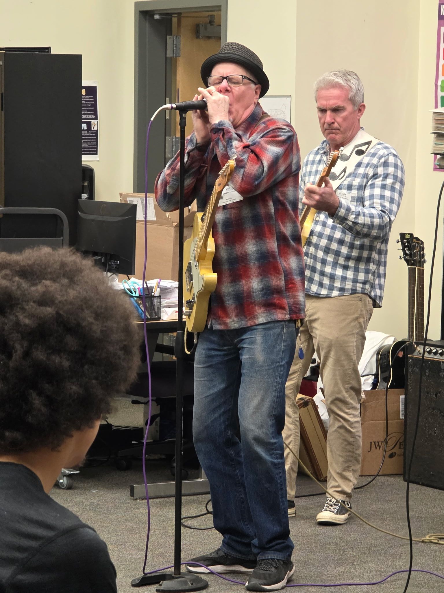 Man singing into a microphone onstage while another man stands behind him with a guitar.