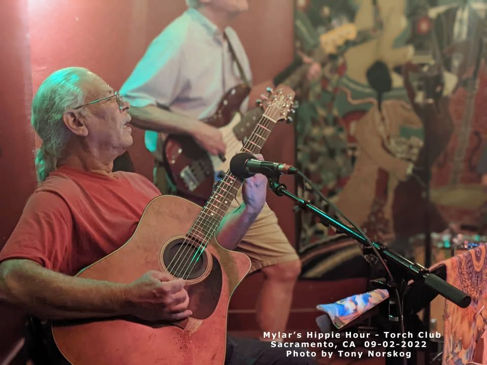 Musician playing guitar on stage, with another performer in the background