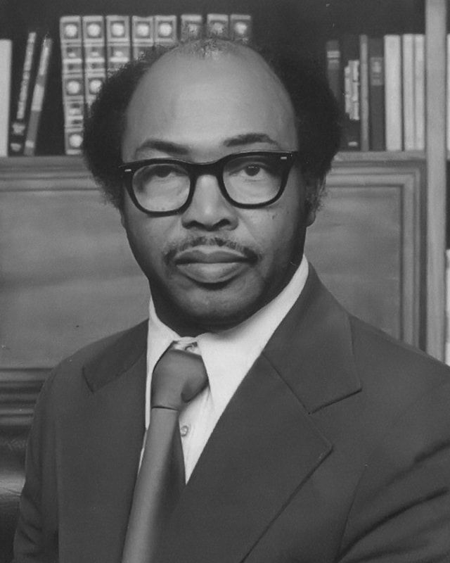 Portrait of a man in a suit and glasses posed before a bookshelf