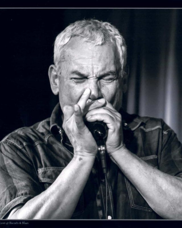 Black-and-white close-up of a man playing harmonica, eyes closed and hands cupped around the instrument.