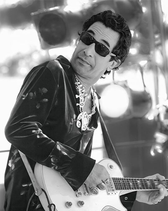Musician in sunglasses playing a white electric guitar on stage under bright lights
