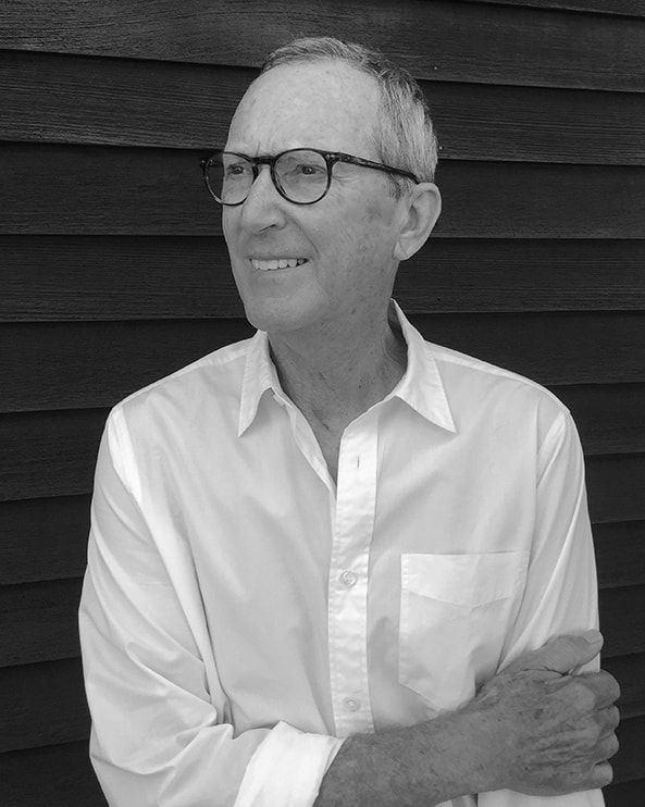 Smiling man in glasses wearing a white button-down shirt, standing against dark siding.
