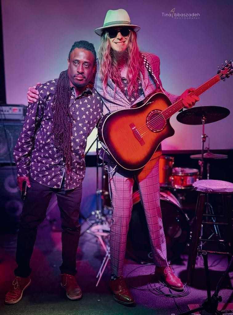 Two musicians pose onstage under purple lighting, one holding an acoustic guitar.